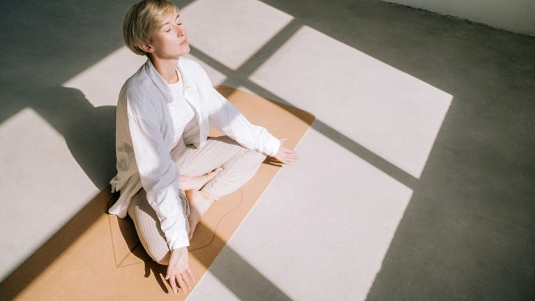 Beautiful authentic woman with short blond hair is meditating sitting in lotus position on yoga mat in front of a window. She is wearing a light-colored casual clothing. Concept of relaxation exercises