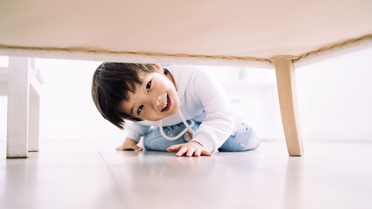 Portrait of smiling child sitting on floor and looking under sofa while playing hide and seek. 