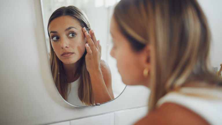 Close up of beautiful young woman looking in the mirror while touching her face and checking for pimples, wrinkles or bags under eyes during her morning beauty routine