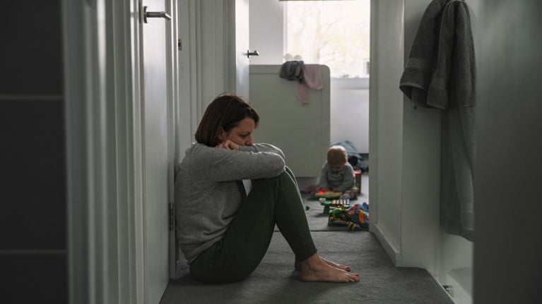 Mature mother sitting on landing floor whilst her baby plays in the background - post natal depression