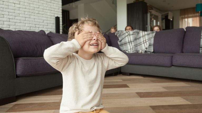 Kid boy playing hide and seek game at home, child closing eyes with hands counting while parents and sister hide behind sofa in living room peeking out, happy family having fun with children concept