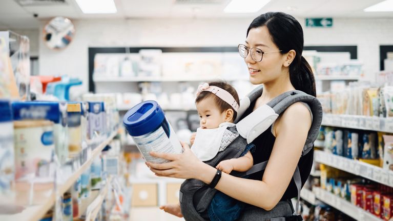Young Asian mother carrying cute baby girl shopping for baby product in a shopping mall and is looking at a variety of baby formula