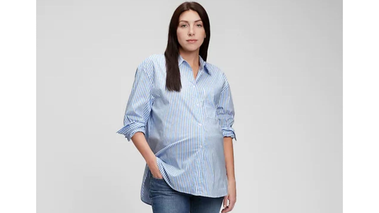 A pregnant woman has her hand in her jeans pocket wearing an oversize blue and white striped button-up shirt