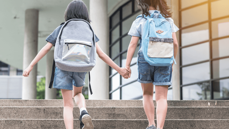 two girls holding hands wearing backpacks walking into school