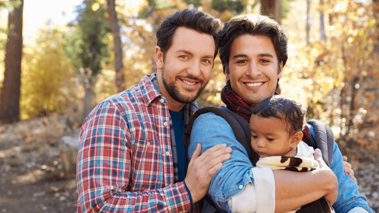 two dads standing in a forest where the leaves are changing holding their baby
