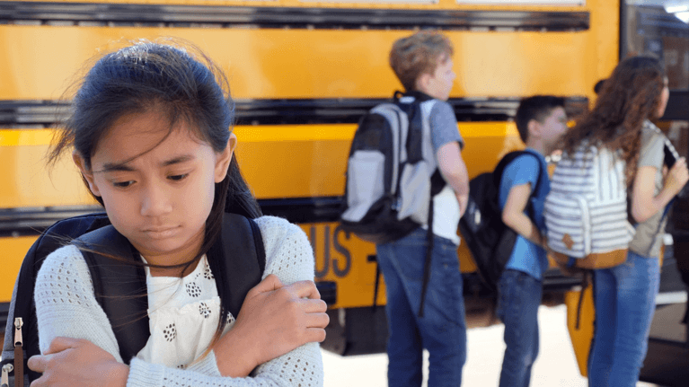 child standing in front of other kids and a school bus and looking sad