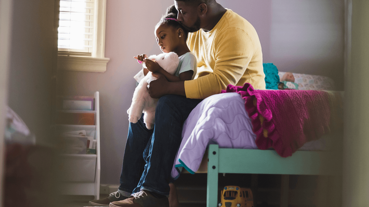 father and daughter sit on a bed reading a book together