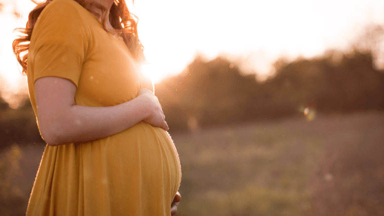 pregnant woman wearing a yellow dress standing in a field holding her stomach 